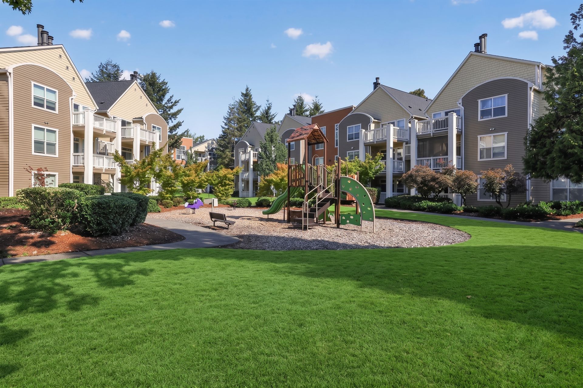 Apartment complex courtyard with green lawn, playground, and landscaped gardens under a blue sky