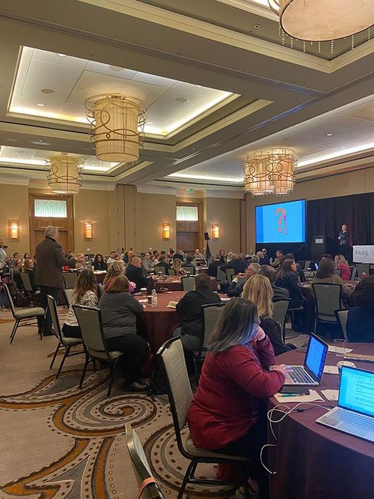 A large room filled with people sitting at tables with laptops.