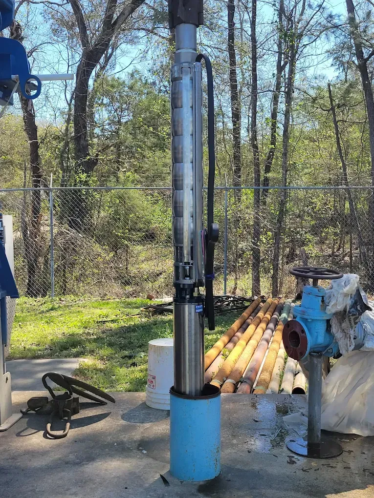 Outdoor drilling rig with tall metal pipe in a wooded worksite, surrounded by stacked orange pipes and equipment