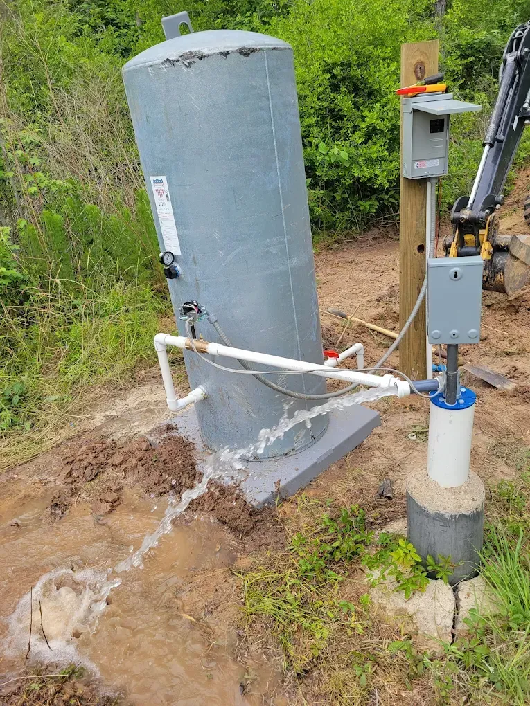 Cylindrical metal tank with white pipes beside an outdoor utility post in a grassy dirt area