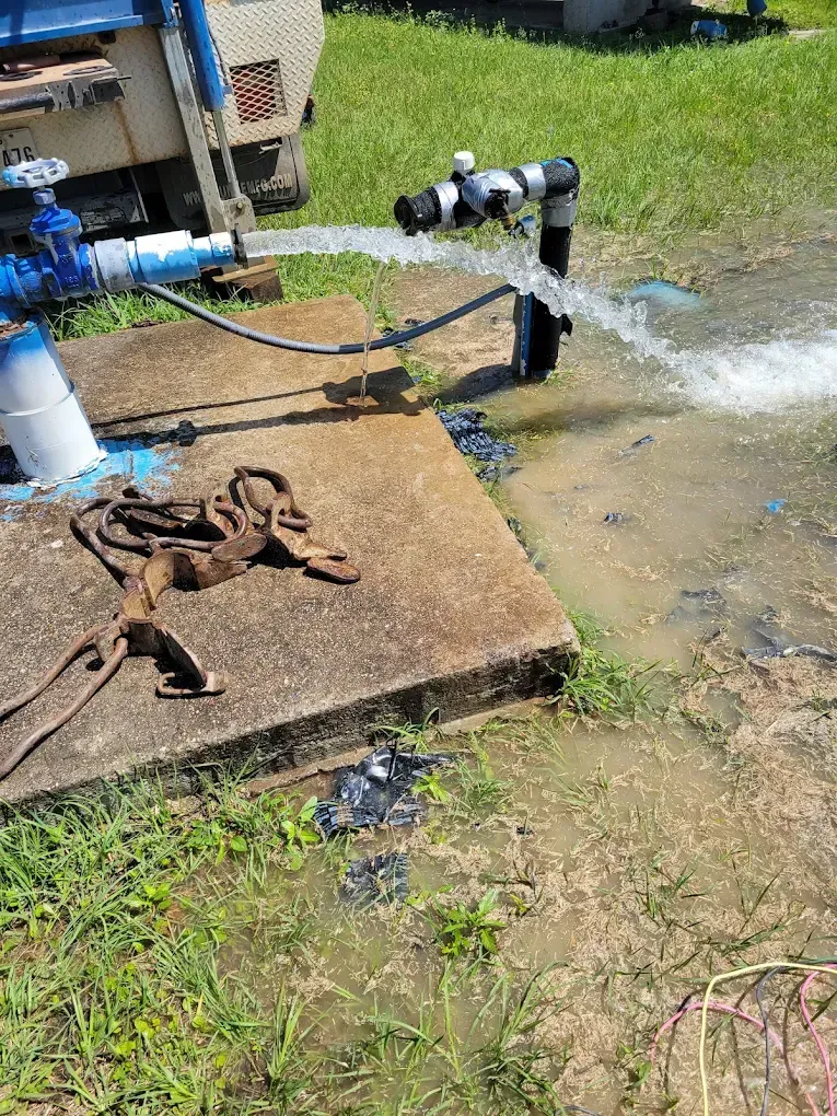 Water pipe spraying into a muddy yard beside a concrete slab and coiled hose