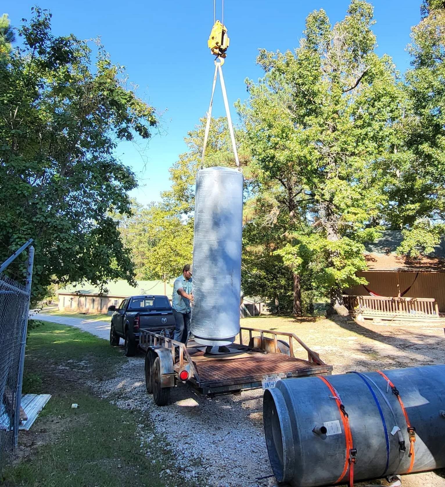 Crane lifting a large gray metal cylinder on a trailer in a sunny yard with trees.