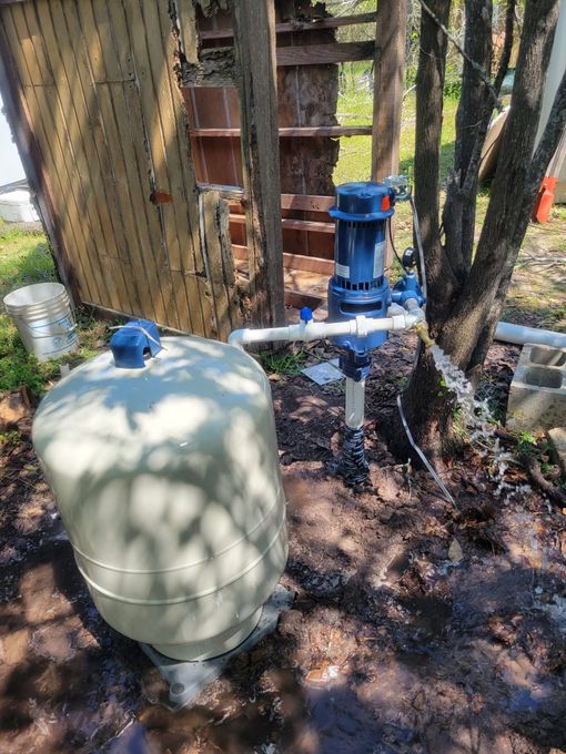 Blue irrigation pump and beige pressure tank beside trees in a wooded yard