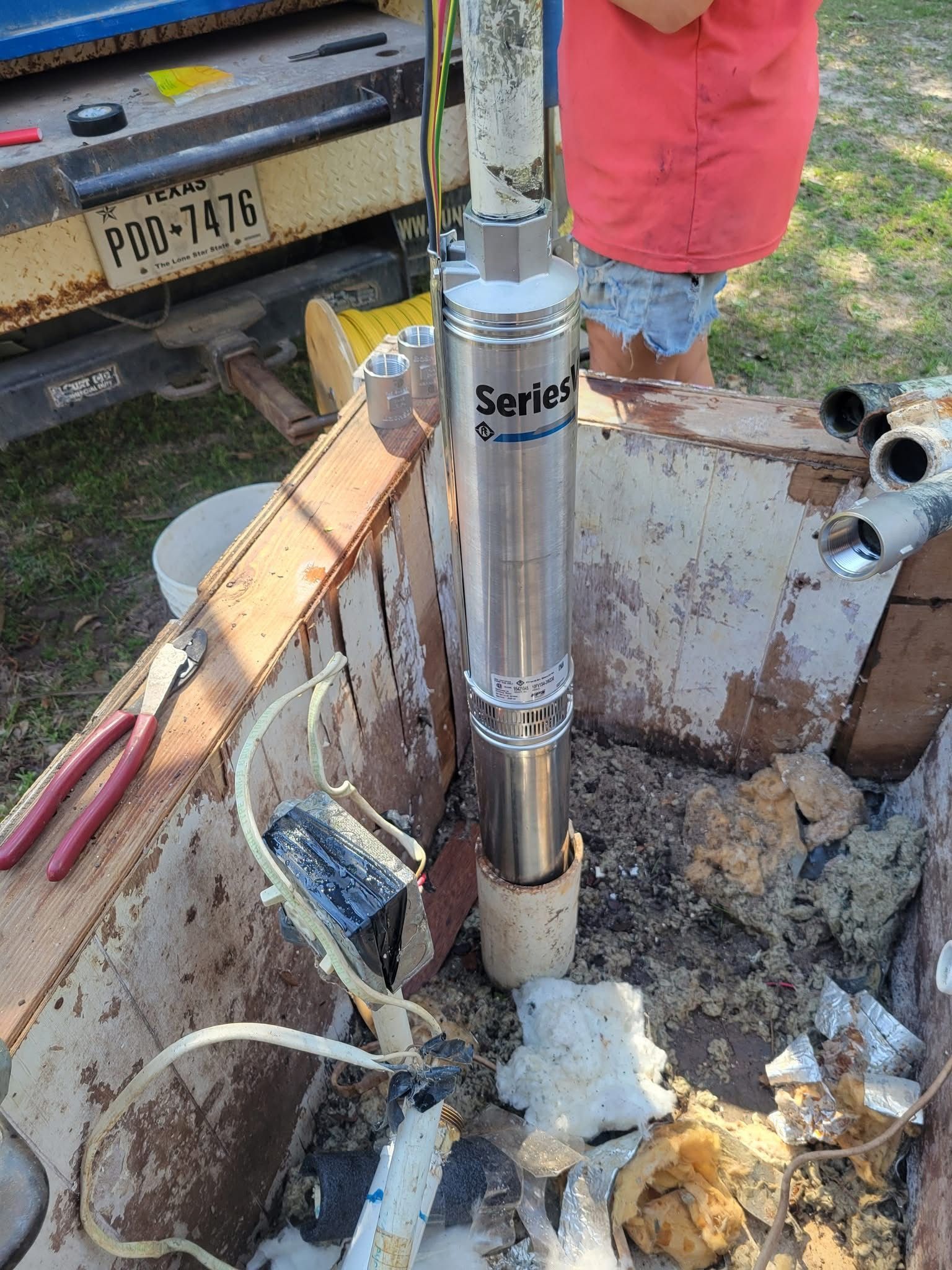 Water well pump being installed in a muddy excavation beside a truck and tools.