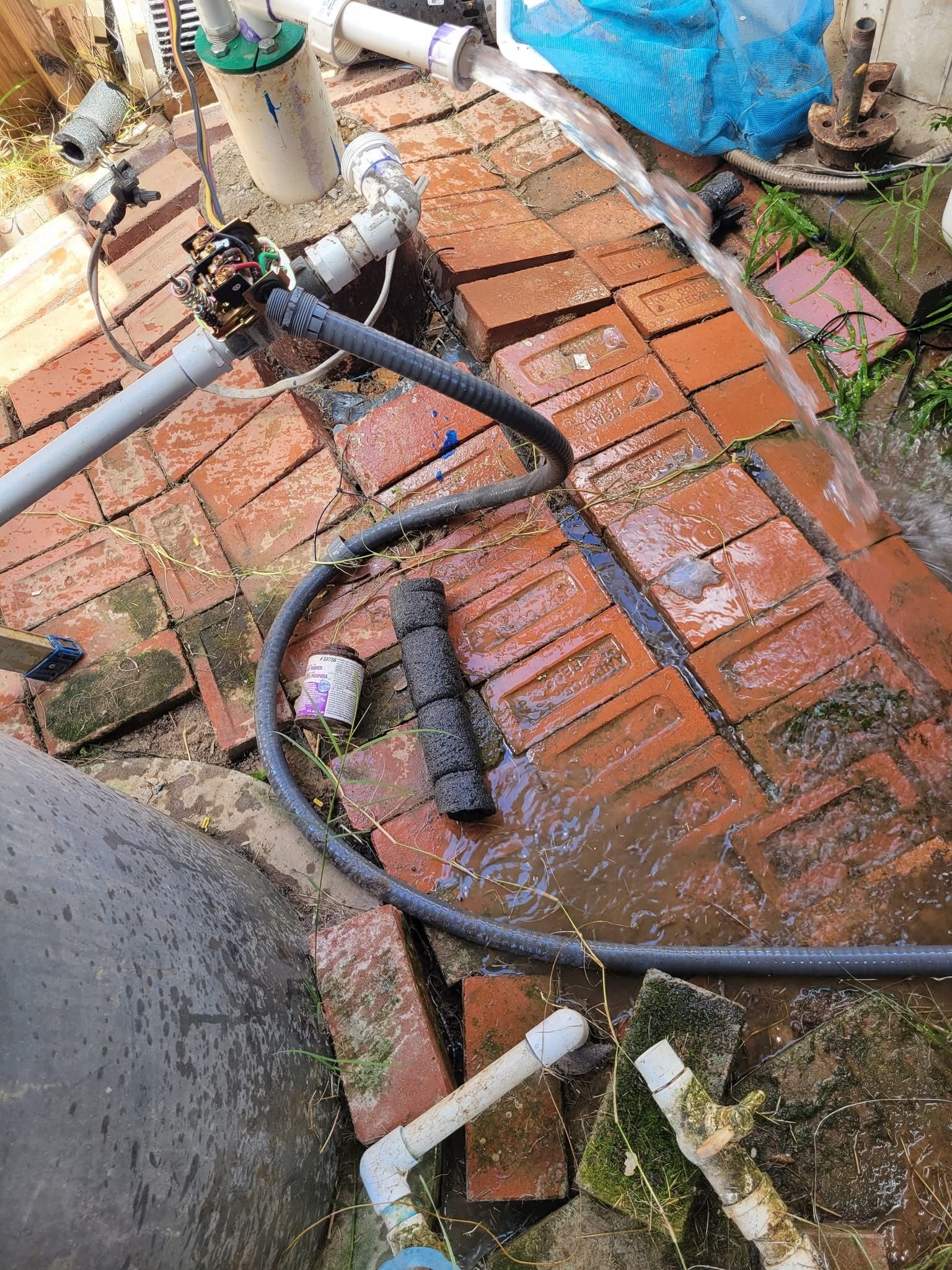 Outdoor brick patio with wet black hoses, white pipes, and a blue tarp near a wall