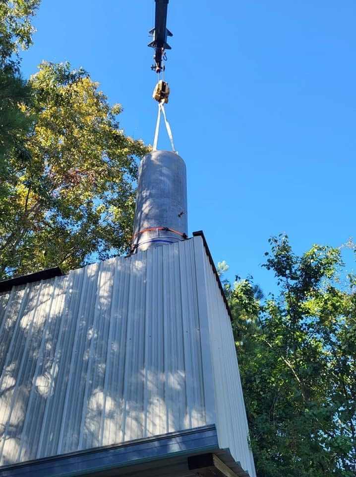 Cylindrical tank being hoisted by crane above a corrugated metal roof under a blue sky