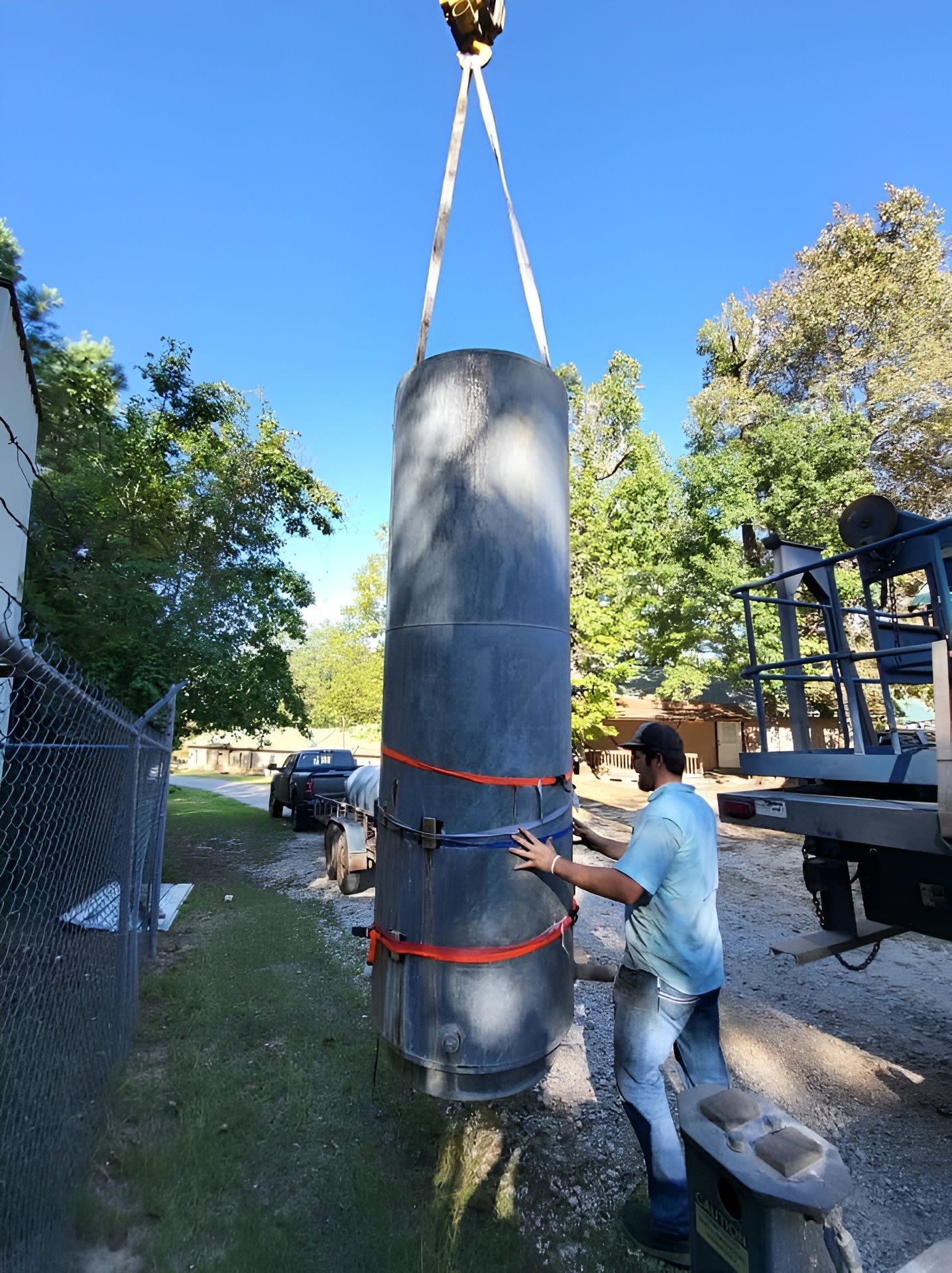 Worker secures a large blue cylinder hanging from a crane in an outdoor yard