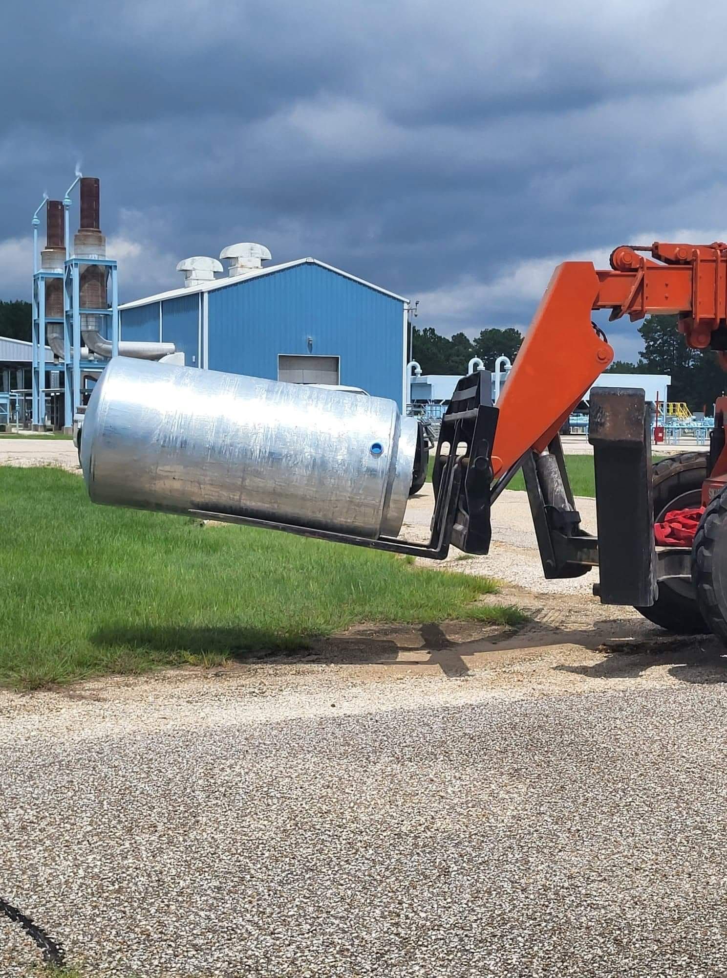Heavy equipment lifting a large shiny metal tank beside a blue industrial building under cloudy skies
