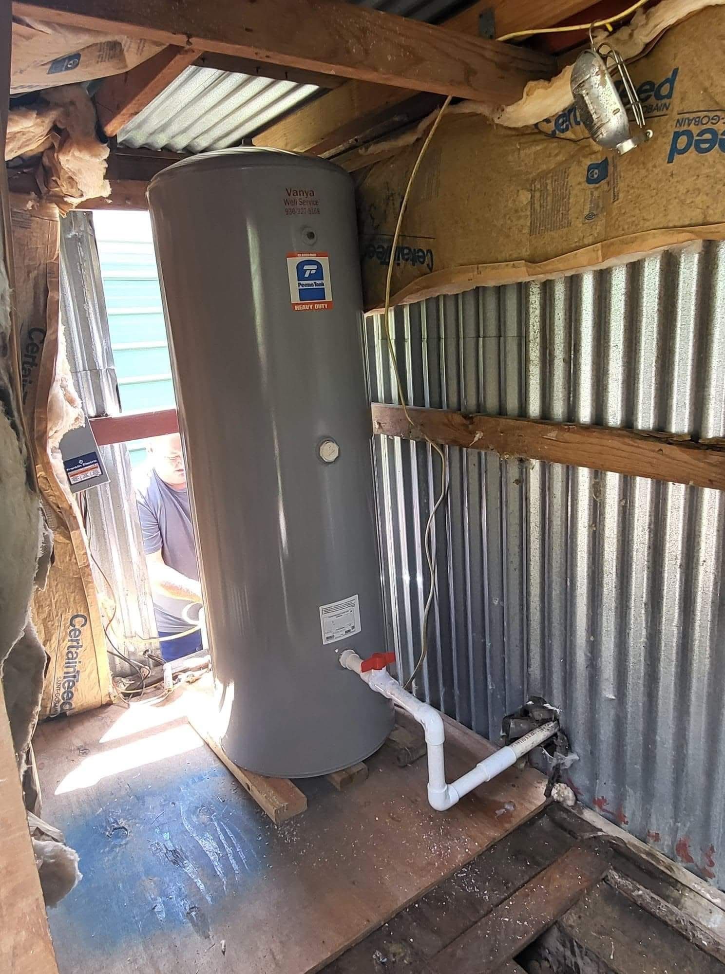 Tall gray water heater in a small corrugated-metal utility room with exposed roof beams and piping.