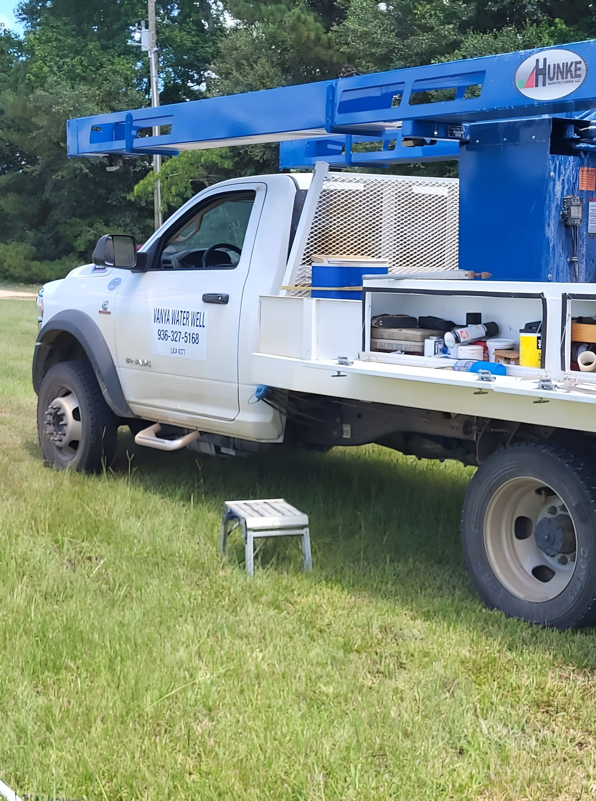 White utility truck with blue ladder lift parked on grass, side compartments open with tools visible.