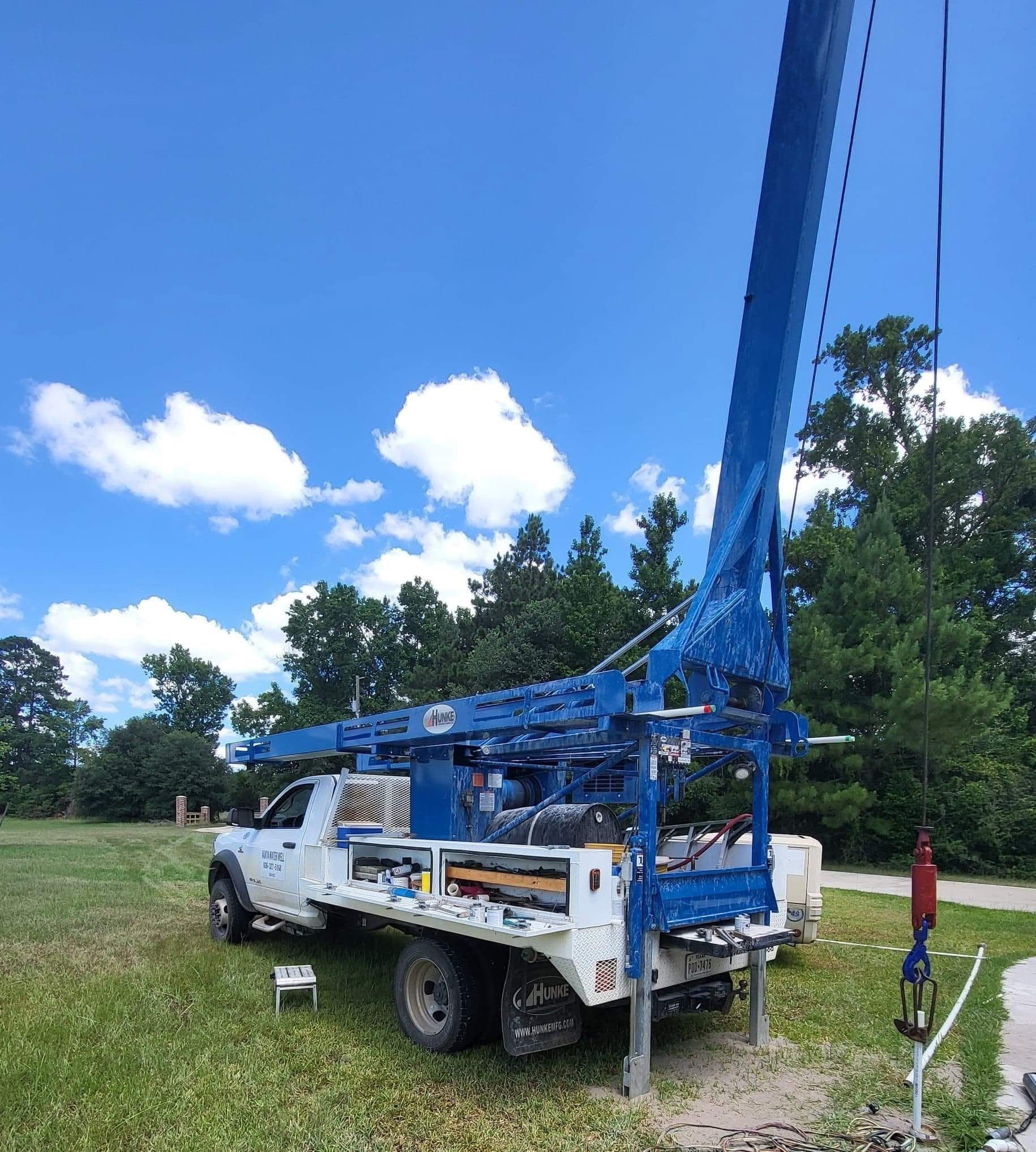 White utility truck with a tall blue drilling rig parked in a grassy field under a blue sky