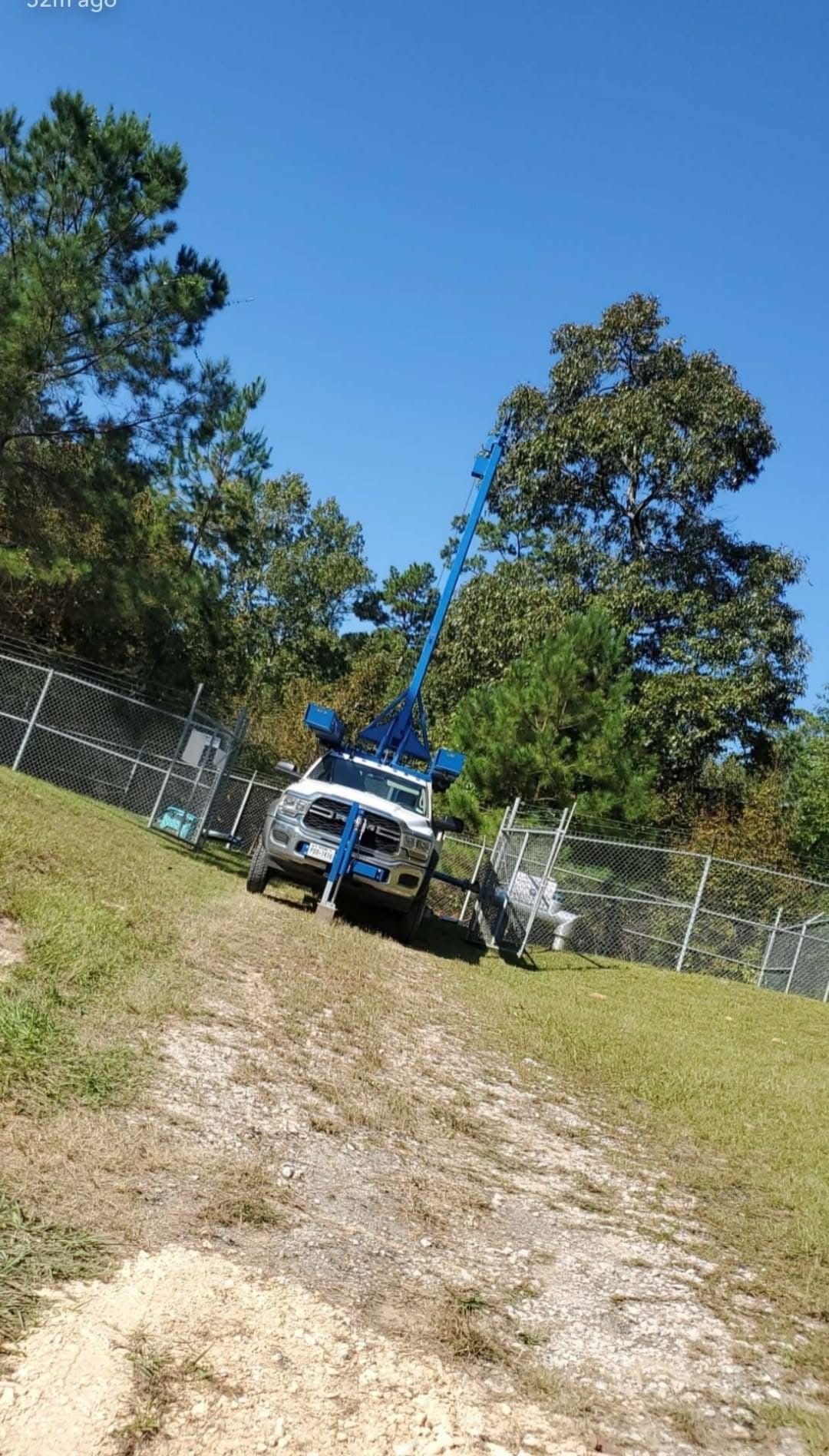 Blue utility truck on a grassy driveway, with trees and chain-link fences under a clear sky.