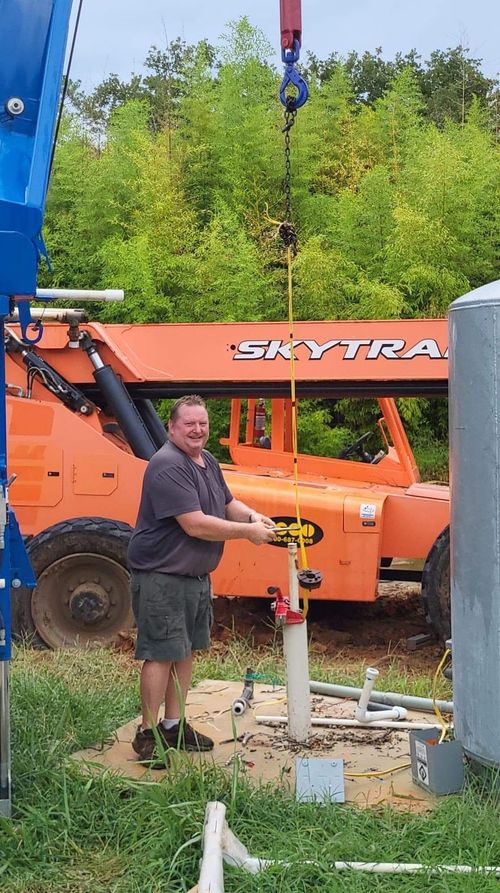 Man standing by a hanging cable and orange boom lift in a grassy outdoor worksite