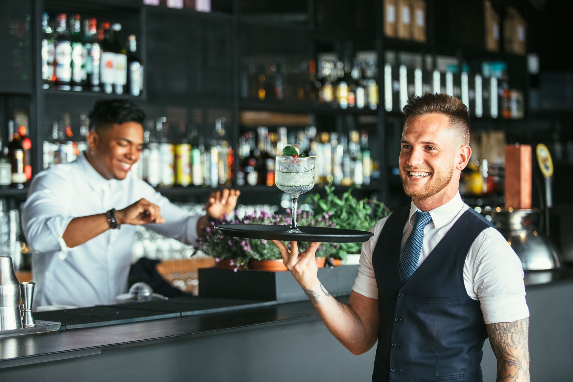 Bartender carrying a cocktail tray in a modern bar, with bottles and glassware behind him.