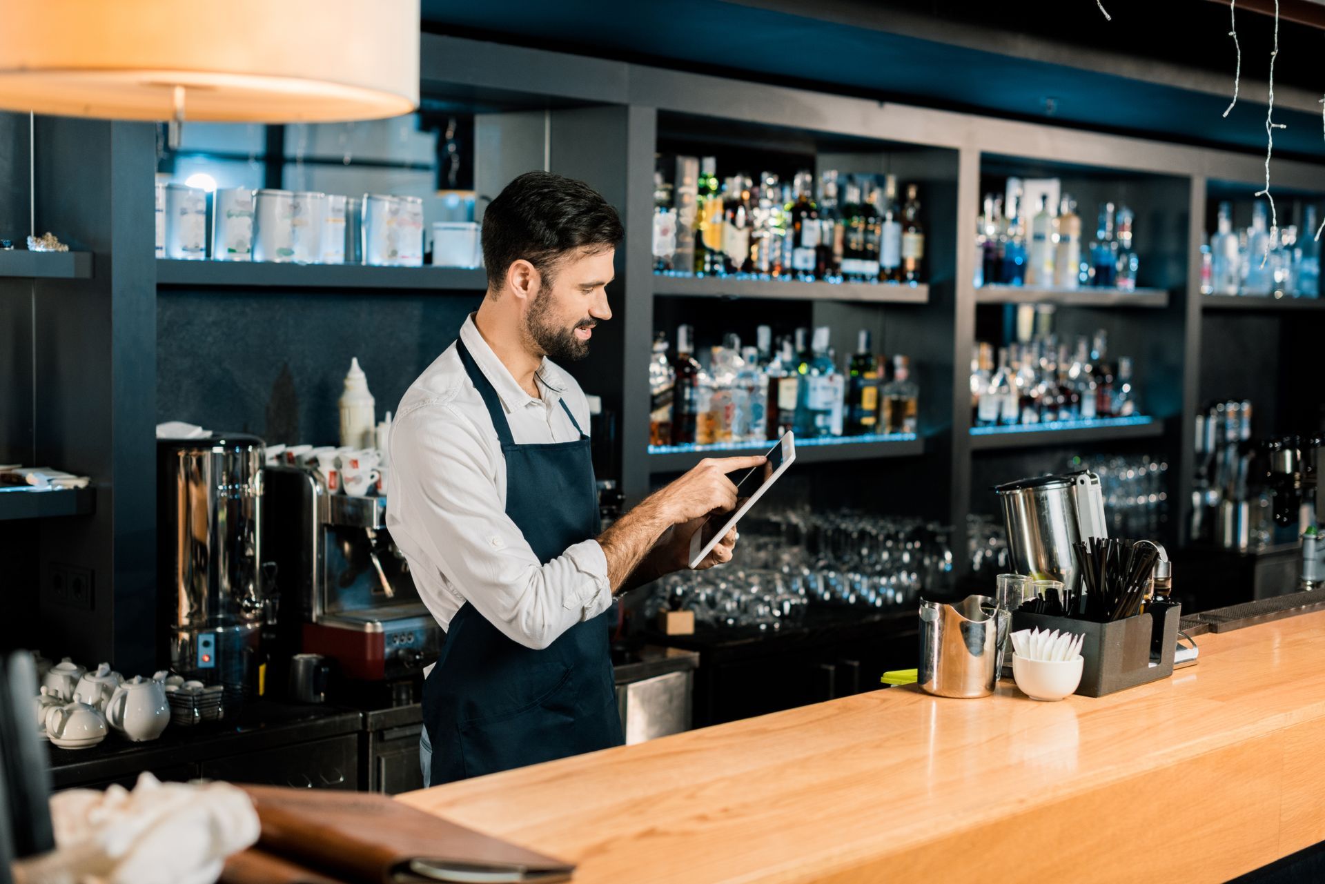 Bartender in apron using a tablet behind a well-stocked bar with glassware and bottles