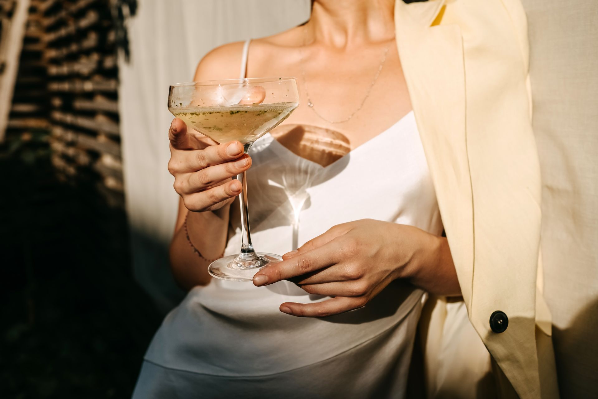 Person in a cream blazer holding a martini glass and small white clutch in warm indoor lighting