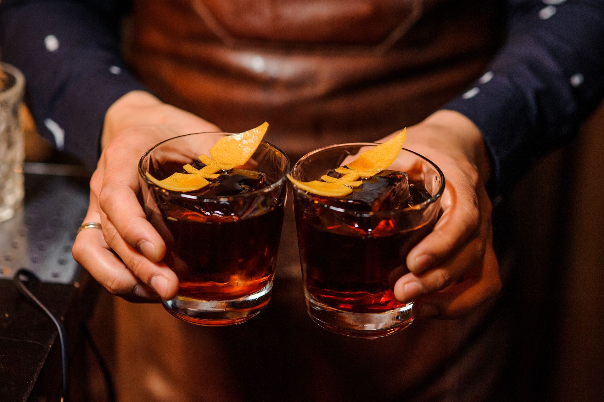 Two hands holding whiskey cocktails with orange peel garnish over a brown apron