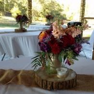 Floral centerpiece with red, pink, and cream flowers on a rustic wood slab at a decorated outdoor event table