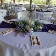 Round banquet table with lavender tablecloth, white chairs, purple napkins, and floral centerpiece under a tent