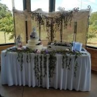 Decorated dessert table with white drape, hanging greenery, and a tiered cake by large windows