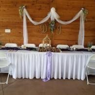 Decorated banquet table with white drapes, lavender accents, and floral centerpieces against a wood wall