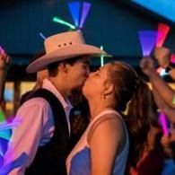 Two people, one in a cowboy hat, kissing at a colorful nighttime party with neon lights