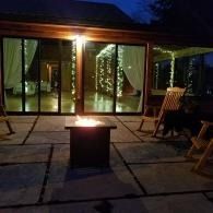 Night patio with a lit fire pit, glass sliding doors, and chairs beside a cozy house exterior.