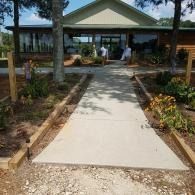 Concrete walkway leading to a small building entrance, bordered by landscaping and trees.