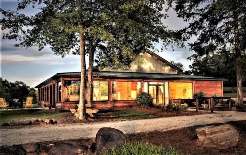 Rustic wooden house at sunset, warmly lit windows under tall trees, with a dirt road and logs in front.