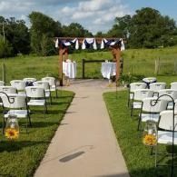 Outdoor wedding aisle with white chairs and lanterns leading to a decorated arch under blue sky