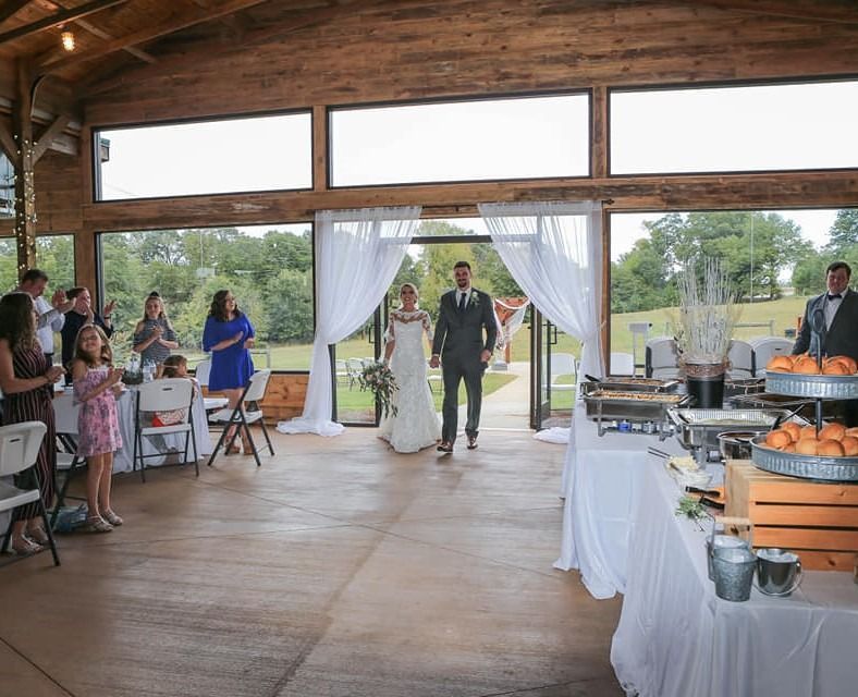 Wedding couple entering rustic reception hall through white drapes, guests seated nearby and buffet set up.