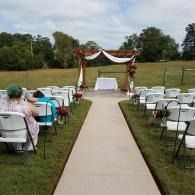 Outdoor wedding aisle with white chairs and a decorated arch under a grassy field and cloudy blue sky backdrop
