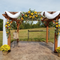 Flower-covered wedding arch with white drapes and yellow blooms on a concrete path overlooking a field