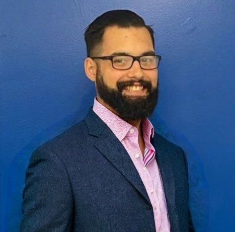 A man with a beard and glasses is wearing a suit and a pink shirt.