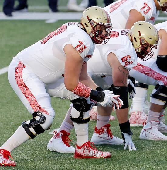 Football players crouched at the line of scrimmage on a field, wearing white uniforms and gold helmets.