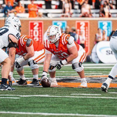 American football line of scrimmage, orange-clad lineman crouched over the ball on the field