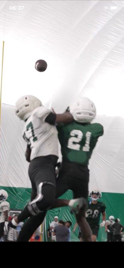 Two football players in white and green jerseys leap for a ball during practice indoors