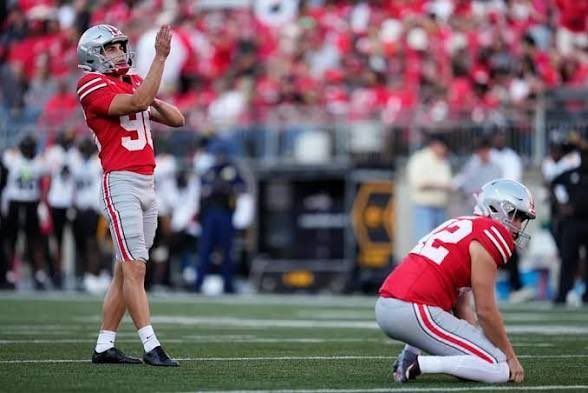 Football player in red kicks as teammate kneels on field during game, crowd blurred in background