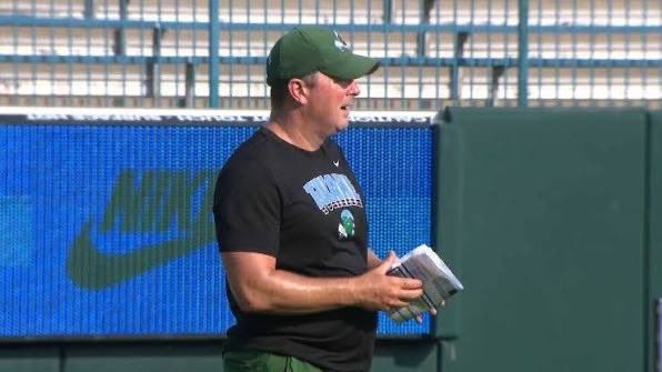 Coach in green cap and black shirt holding a notebook on a sports field with a blue Nike sign in the background