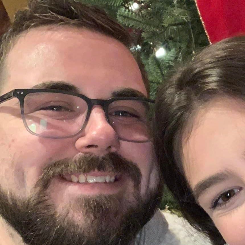 A man with a beard and glasses is smiling next to a woman in front of a christmas tree.