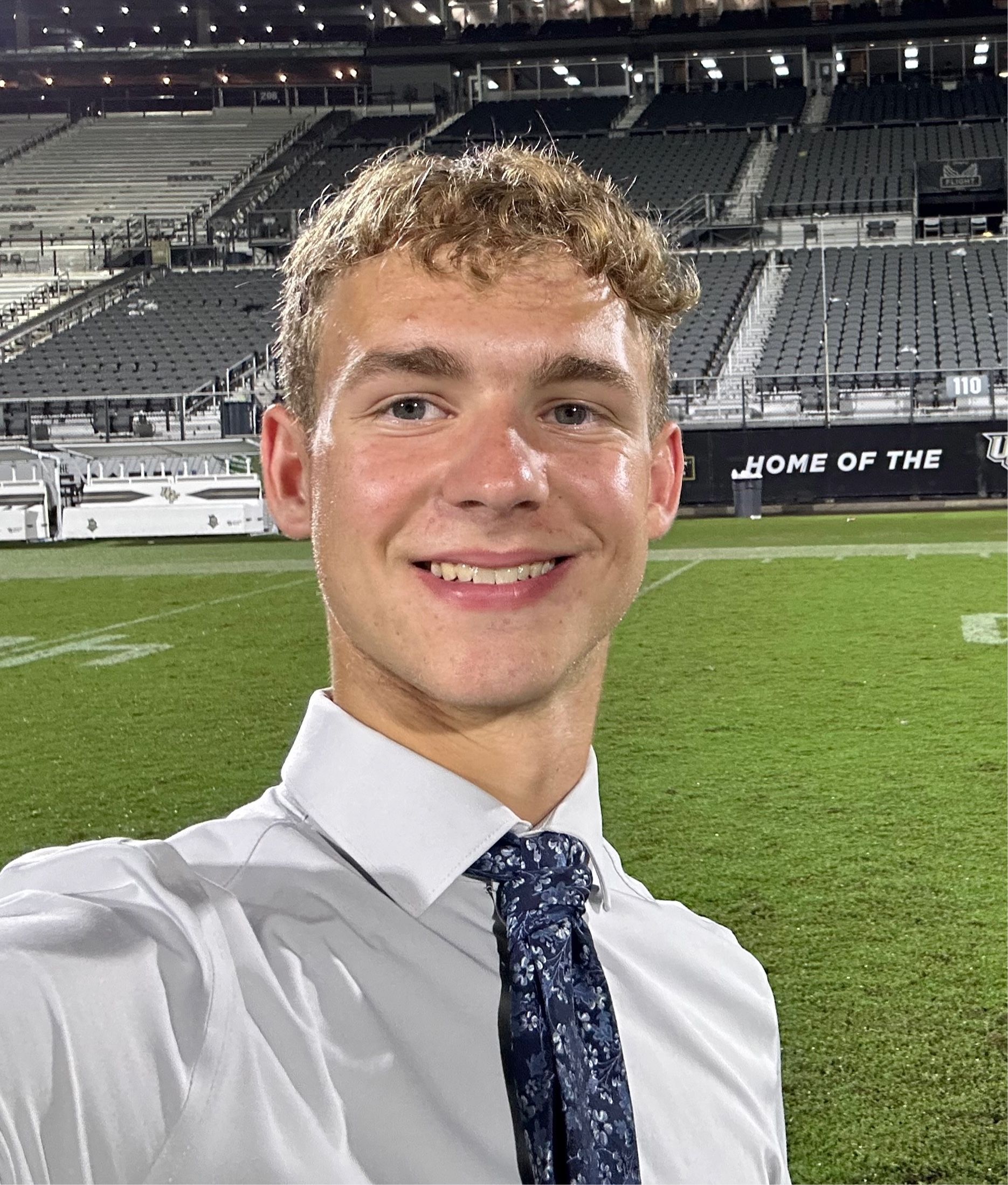 Selfie of a smiling man in a white shirt and blue tie at a mostly empty stadium at night