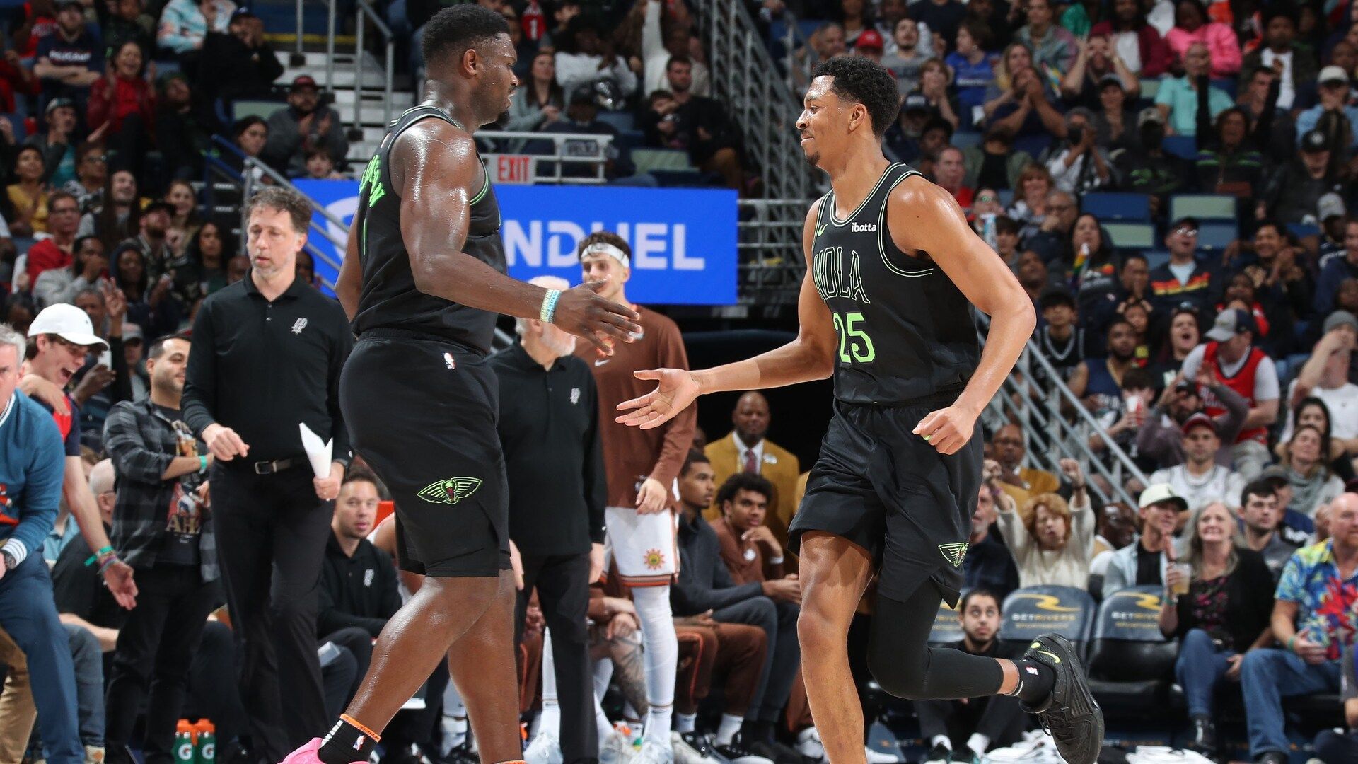 Basketball players in black and green jerseys exchange a handshake during a game with cheering fans behind them