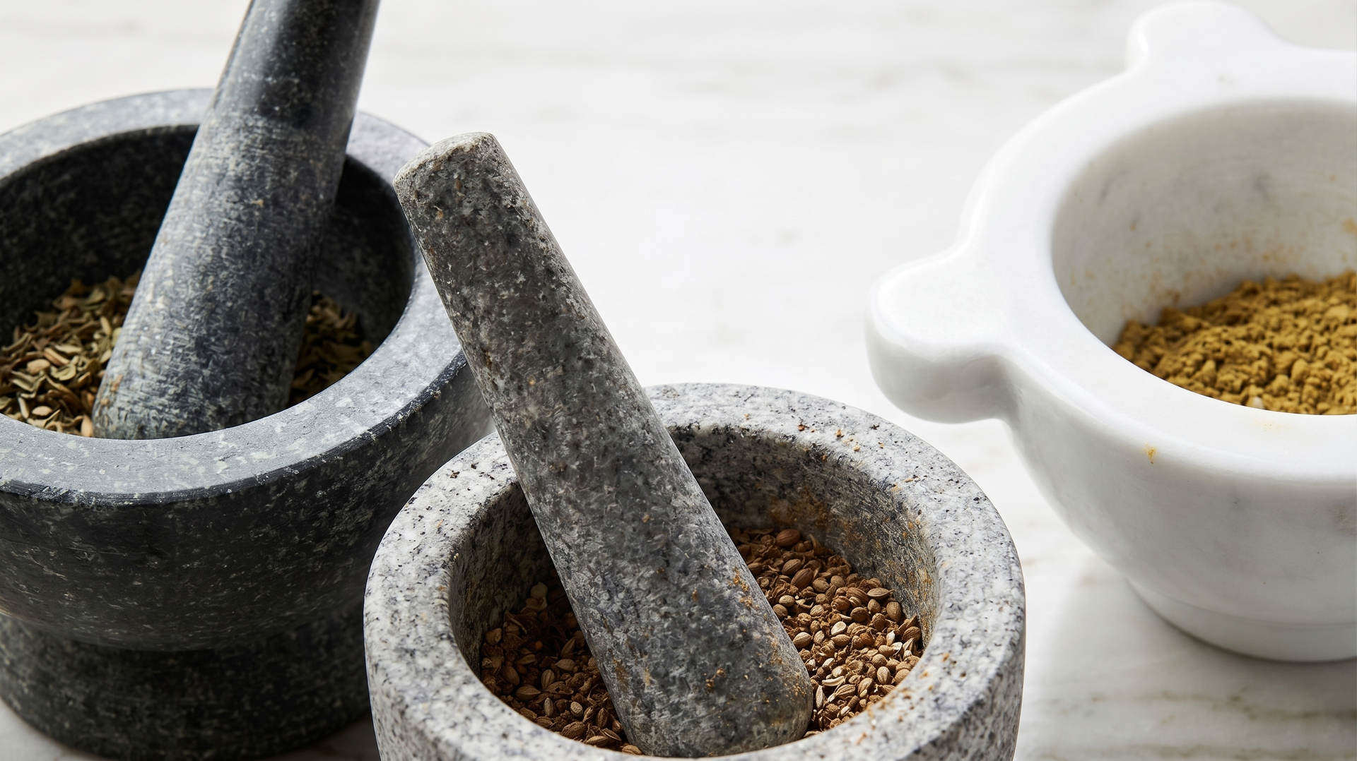 Two stone mortars and pestles with ground spices, beside a white bowl on a light surface.