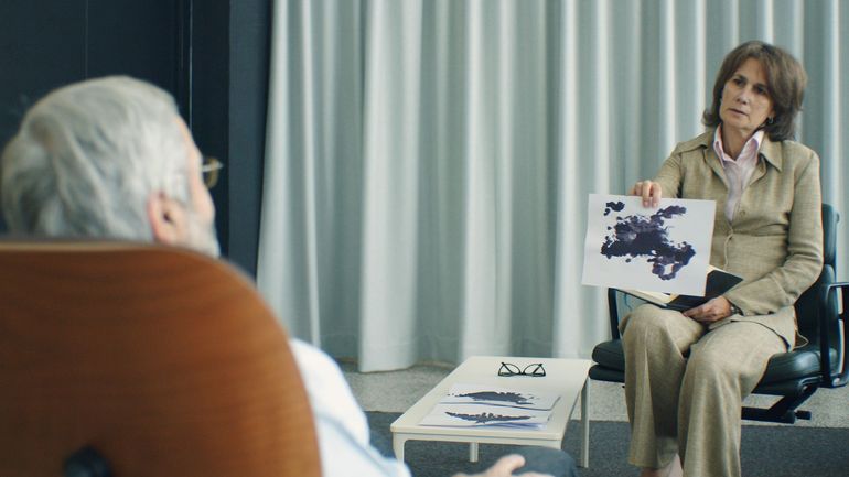 A professional holding a Rorschach inkblot test card across from a patient in a clinical room.