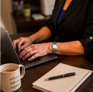 Therapist typing on a laptop at a desk with a notebook, pen, and coffee mug nearby.