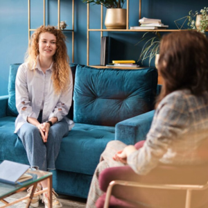 Two people sit on a black couch in a bright office during a consultation, while an unseen person takes notes.