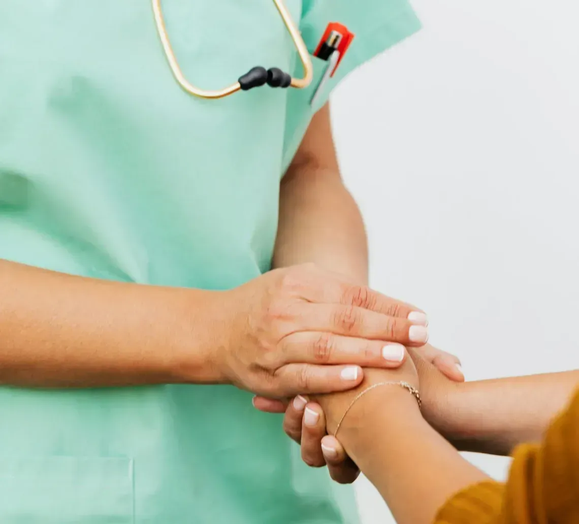 A healthcare worker in green scrubs holds a person's hands in a comforting gesture, with a stethoscope around their neck.