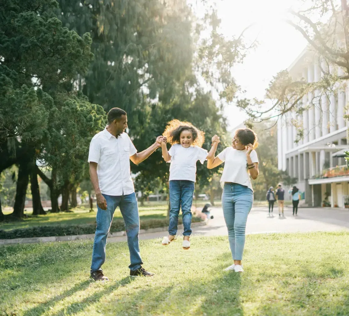 A family holds their child’s hands while lifting them into the air in a park on a sunny day.