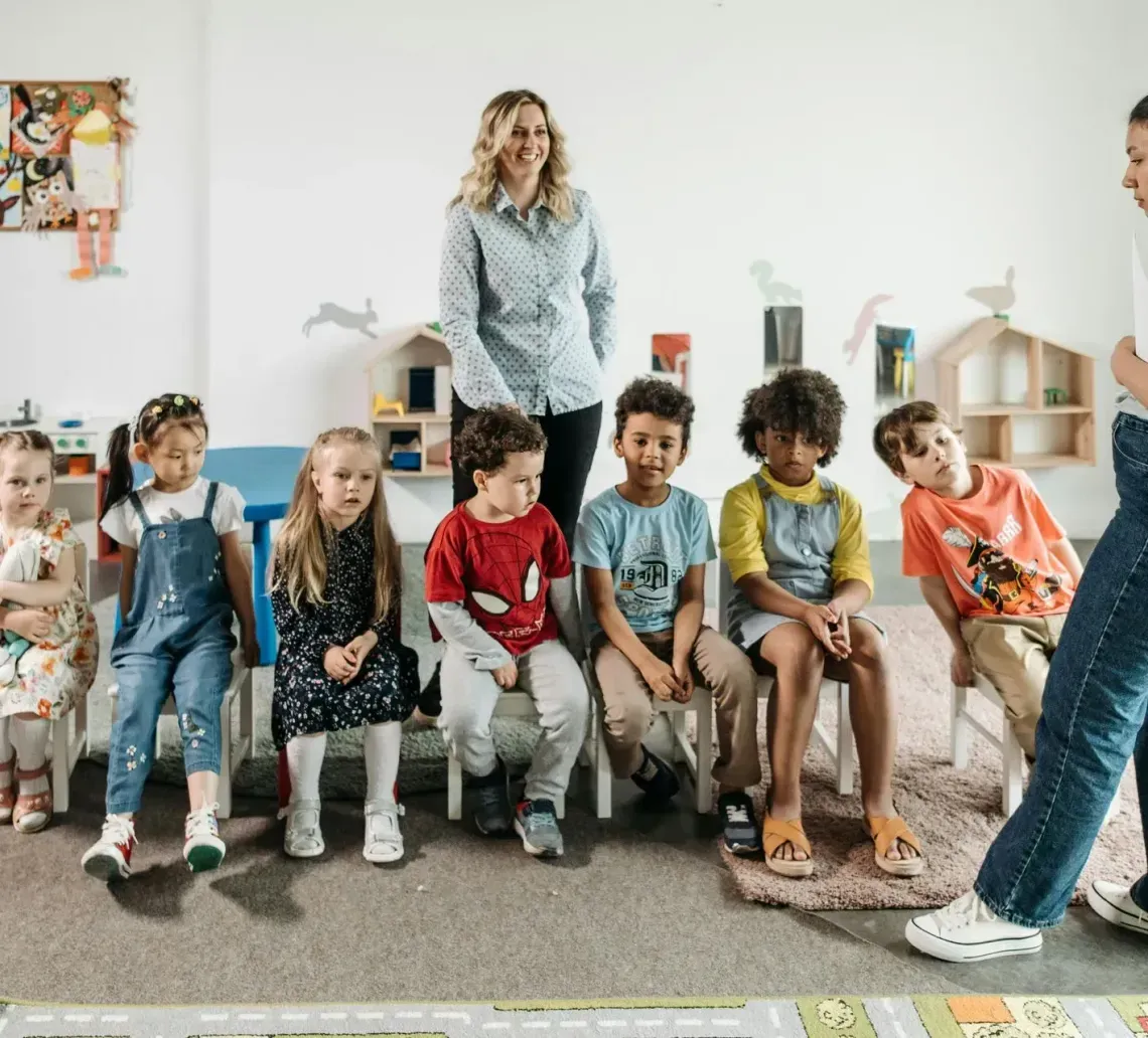 A teacher stands before a group of children seated in a row on chairs in a brightly lit, decorated classroom.