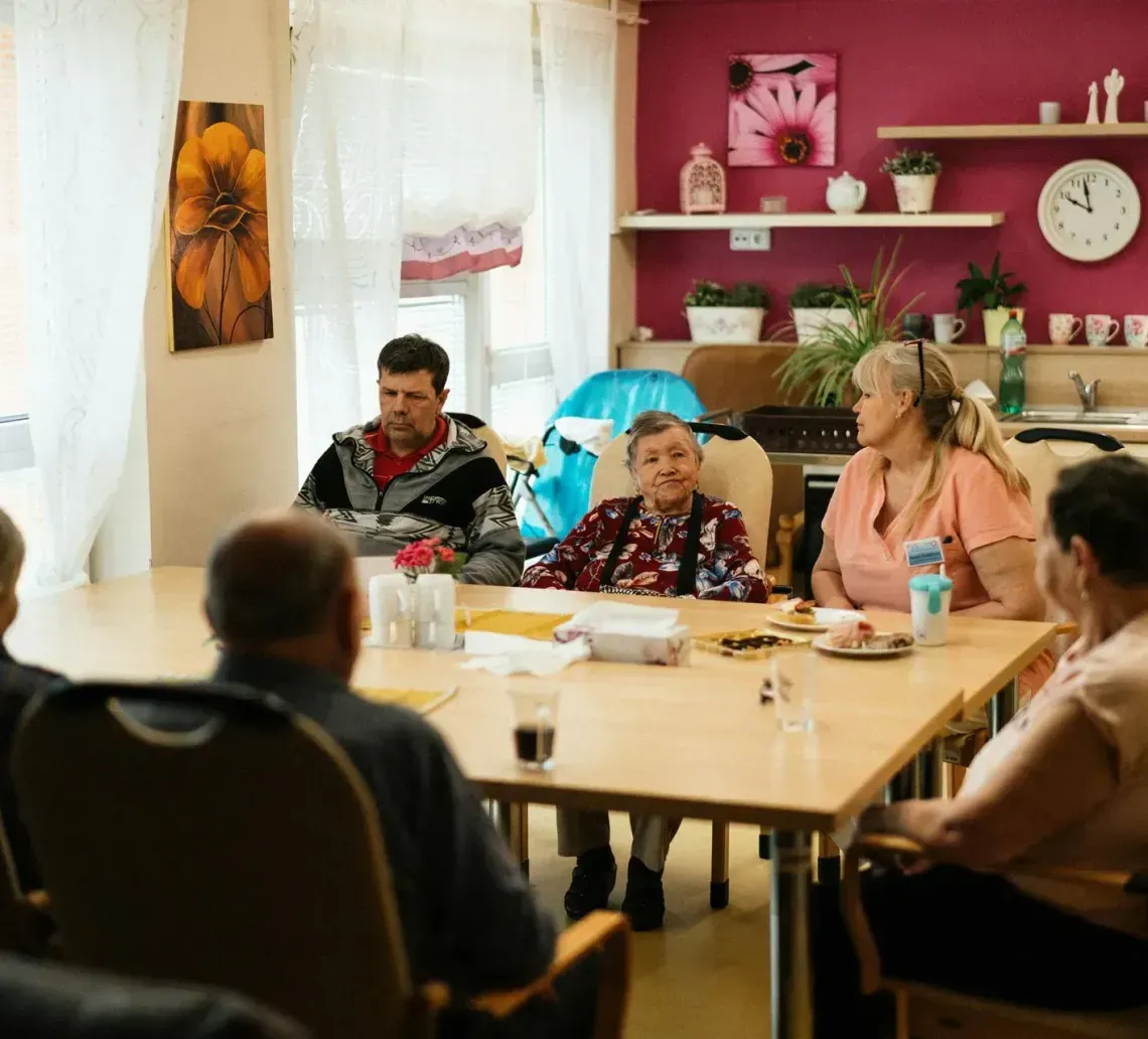 People sit around a dining table in a bright, community-style room with a red accent wall, sharing a social moment.